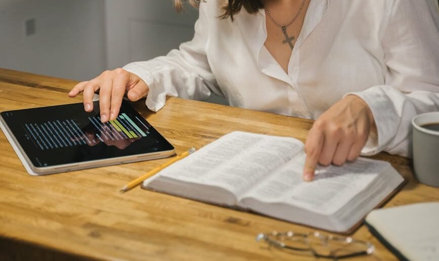 A woman studies the Bible at a wooden table, combining printed and digital texts.
