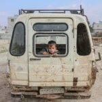 A young boy peers from an abandoned van in the desolate streets of Idlib, Syria, amidst war devastation.