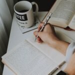 Person writing in a notebook on a desk with a coffee mug, creating a cozy workspace vibe.