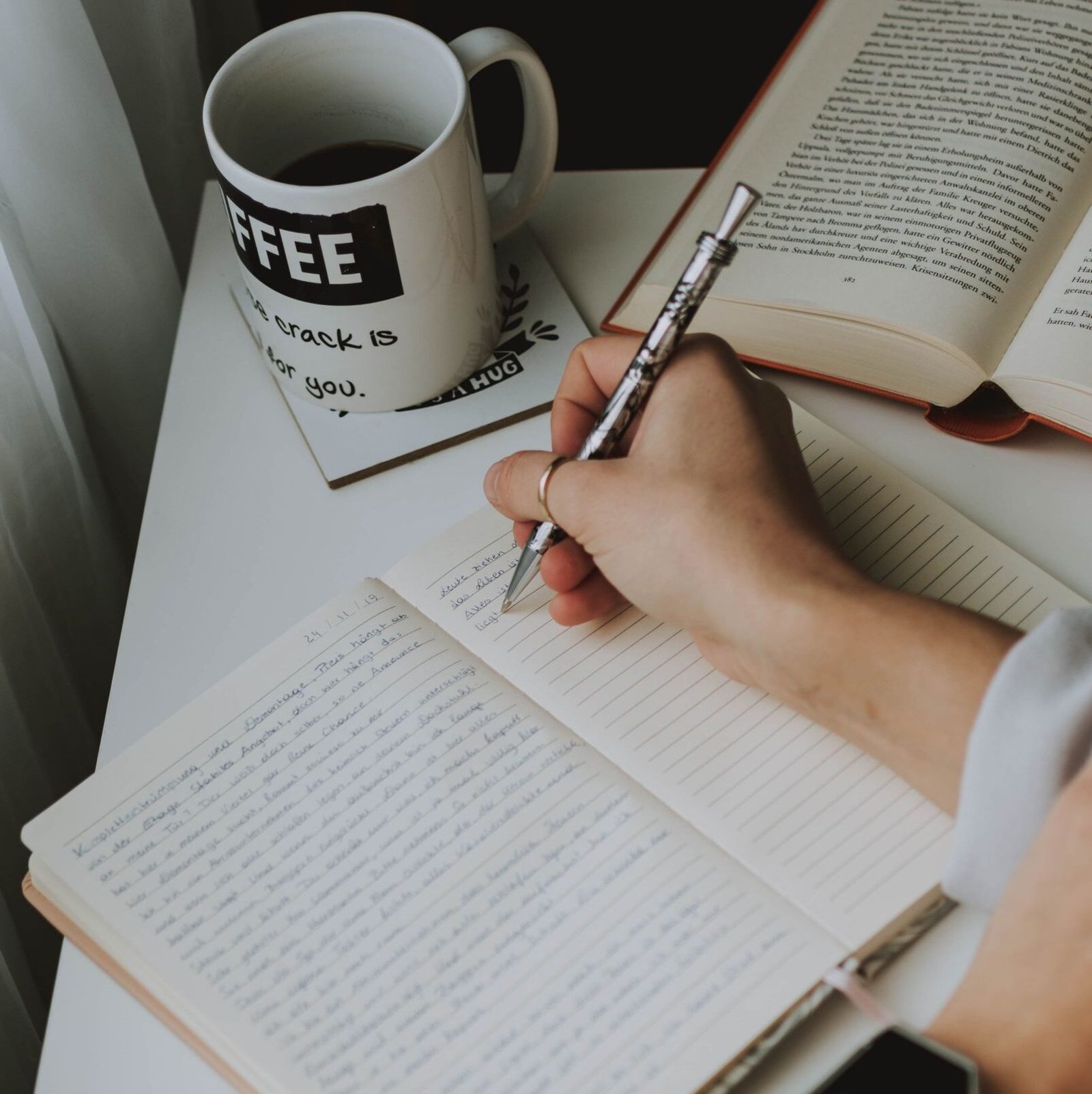 Person writing in a notebook on a desk with a coffee mug, creating a cozy workspace vibe.