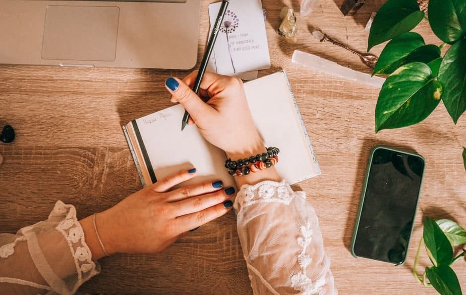 Hands writing in a notebook on a wooden desk with plants, laptop, and phone nearby.